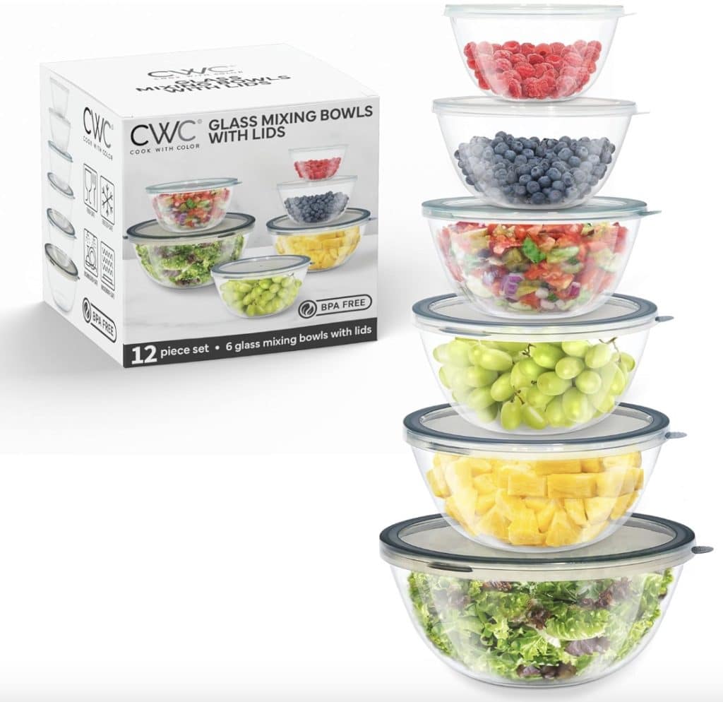 A box labeled "Glass Mixing Bowls with Lids" sits beside a stack of six glass bowls filled with an array of foods: raspberries, blueberries, green and red grapes, pineapple chunks, and salad. Each bowl has a matching lid—perfect for storing your favorite baked apple fritter recipe ingredients.